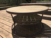 Photo of a round outdoor table showing metal frame and wooden slat top with metal grid shelf underneath on wooden deck surface in daylight.