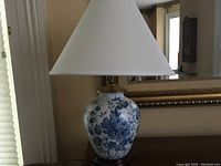 Full view of the porcelain table lamp with white conical shade on a wooden surface in front of a mirror.