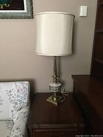 Full view of brass-tone table lamp with cream-colored drum shade placed on brown wooden side table near floral upholstery chair.