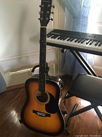 Steel string acoustic guitar shown with sunburst finish, resting on a black adjustable guitar stand near a keyboard and chair on hardwood floor.
