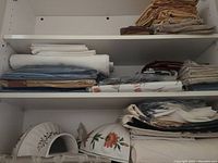 Shelf with stacks of folded vintage tablecloths, napkins, and linens along with two ceramic food covers with floral designs on lower shelf.