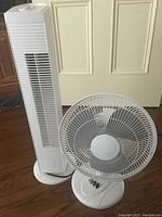 Photo showing the white tower fan and the white oscillating desk fan side by side on hardwood floor in front of a door.