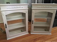 Photo showing two white vintage wall-mounted curio cabinets with glass doors and decorative carved molding, placed on wooden floor.