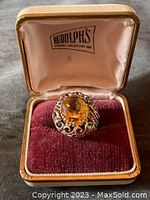 Front view of ring in Rudolph’s jewelry box showing oval yellow gemstone and filigree setting