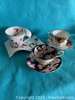 Three tea cup and saucer sets arranged on turquoise background: black floral, rose band, tulip with square saucer