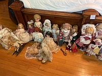 Wide view showing a row of antique dolls seated against a wooden headboard, highlighting a variety of styles, clothing, and materials.