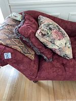 Side angle showing the red fabric sofa with curved rolled arm, floral upholstery, and multiple decorative pillows on top