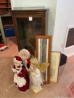 Wooden display cabinet with glass door and Christmas-themed decor in front along with framed wall art pieces leaning against the cabinet