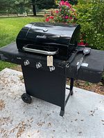 Front view of black Char-Broil gas grill with closed lid, two control knobs, side burner, wheels, and included brushes on right shelf.