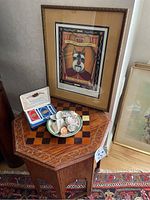 Photo showing octagonal wooden chess table with carved floral details topped with framed dog artwork, a bowl of seashells, and a tin box holding playing cards.