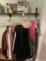Photo of multiple winter coats hanging on rack including pink, brown, black, and green colors, with black gloves, pink hat, and green scarf. Paper and household products are on shelf above.