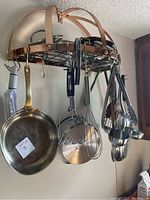 View of metal hanging rack with pans and various kitchen utensils hanging including whisks, ladles, and tongs
