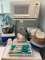 Photo showing microwave, coffee maker, silverware with turquoise handles in white tray, four white coffee mugs stacked, coffee filters in basket, blue cooler, and background oven mitt.