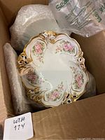 Photo showing ornate porcelain plate decorated with pink roses and gold trim, inside packing box with bubble wrap.