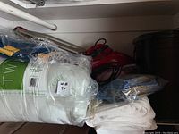 Photo showing several pillows, towels, folded linen cloths, a red Scorpion dust buster vacuum, and some blankets stacked in a closet area.