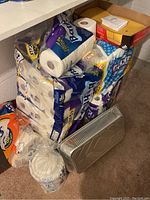 Photo showing multiple packages of paper towels (Brawny) and toilet paper (Scott), disposable paper bowls in plastic wrapping, a box containing empty envelopes, and several stackable metal trays with lids on a carpeted floor beneath a table.