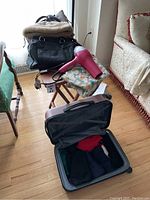 Photo showing an open gray hard-shell suitcase on the floor with women's clothes inside, and a small wooden folding table next to it holding several purses and a red and blue hair dryer.