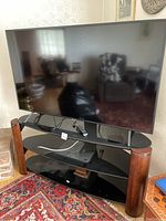Flat screen TV on top of the black glass TV stand with wooden legs. Remote control and cables visible on stand's top shelf.
