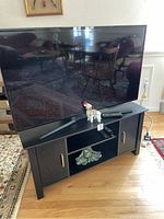 Television and black TV stand with a green frog figurine placed inside the center open shelf of the stand, and a colorful llama figurine on top beside the TV remote.