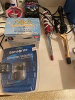 Photo showing two curling irons, Nadin Hair Removal Gel box, and sealed Samsonite converter/adaptor kit arranged on a table.