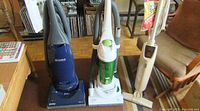 Front view of three vacuum cleaners: Kenmore blue upright vacuum on the left, Eureka white and green upright vacuum in the middle, Hoover white electric broom on the right.