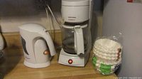 White Black and Decker VersaBrew coffee maker with glass carafe, white electric kettle with orange power button glowing, and packs of circular coffee filters on wooden counter.