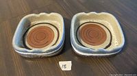 Pair of square glazed earthenware ashtrays with concentric brown circle design, photographed on a dark wood surface with a small numbered tag (18).