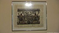 Full view of the hand-painted Egyptian scene on papyrus, mounted in a simple metal frame with glass, hanging on a wall.