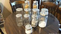 Eleven clear glass food storage containers of various sizes with matching white sealing lids arranged on a dark wood table.
