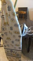 Ironing board covered with fabric patterned with various kitchen-themed illustrations, leaning against a chair. On the chair, there is a white and black Proctor-Silex steam iron with a power cord.