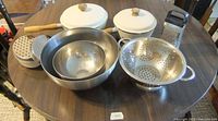 Photo shows two cream-colored thick aluminum enameled pots with lids on the left side, two stainless steel mixing bowls nested on the table, a large stainless steel colander with handles, and two metal cheese graters, one attached to a container and another with two different grate surfaces.