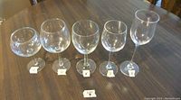 Photo showing 5 different types of clear glass wine glasses arranged in a row on a wooden table. Each glass type has a label with the quantity: 6, 5, 3, 5, 8.