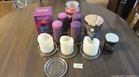 Photo shows a variety of large candles and candle bases arranged on a wooden table. There are three white pillar candles on glass bases, three purple cylindrical candles, one boxed pink candle, one glass jar candle, and one small green jar candle.