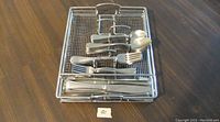 Stainless steel flatware arranged in a stainless steel mesh basket showing knives, forks, and spoons.