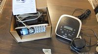 Box containing Uniden cordless phone handset with attached telephone line cord and charging base. Separate NexXtech digital answering machine with power adapter, control buttons, and LCD screen displayed on a wooden surface.