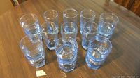 16 clear glass drinking glasses displayed in two rows on a wooden table, 6 full-size and 10 half-size glasses, all matching and in very good condition