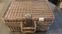 Closed view of a rectangular wicker picnic basket displaying the leather locking strap with brass fittings and wicker handle on the front side.