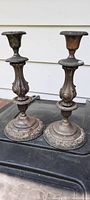Front sideways view showing full height and detail on two antique heavy metal candlestick holders with ornate exterior design and aged finish.