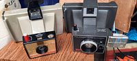 Side angle showing both cameras featuring Polaroid Land Camera and Big Swinger camera on a wood surface with flash attachment.