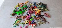 Overview photo showing a large assortment of small, colorful hard plastic toy dinosaurs spread out on a surface.