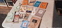 Wide photo showing 16 vintage recipe booklets with various illustrated covers arranged on a white table.