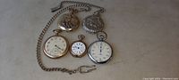 Lot of five pocket watches on beige surface showing two with attached metal chains, including one with hook clasp. Watches vary in size and dial detail, two with stopwatch dials.