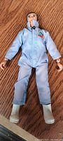 Full view of the small man action figure laying on a wood surface showing blue jumpsuit with A J patch and white boots