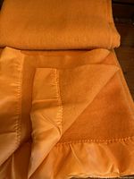 Close-up of the folded tangerine wool blanket showing satin binding and embroidery on edge.