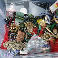 Top-down photo showing mix of green carved beads, gold-tone earrings with pearl centers, metal chains, red tassels, and assorted charms in a plastic container.