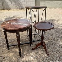 Three small vintage tables displayed together on gravel surface outdoors: Bombay Company wooden piecrust table, round marble top table, and metal framed plant stand with wooden top.