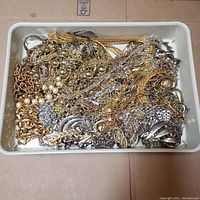 Overview of mixed costume jewelry chains loosely arranged in a shallow white tray, showing various metallic finishes and chain styles.