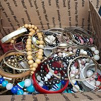 Box filled with a large assortment of vintage bracelets and necklaces, showing various bead types, bangles, and mixed materials spilling over and densely packed