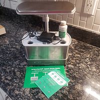 Photo showing the Aerogrow Harvest Elite indoor garden unit on a kitchen counter, including the adjustable LED grow light, power adapter, liquid plant food bottle, and printed setup guides.