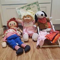 Photo showing four dolls on floor including Cabbage Patch doll with certificate, Raggedy Ann and Andy dolls, and Snoopy plush sitting in chair with extra clothes.